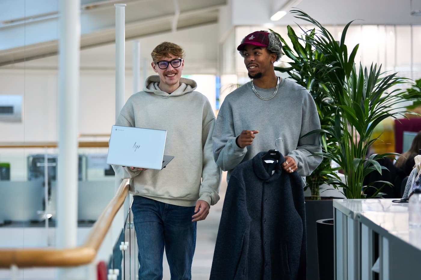 Two men walking indoors and talking, one holding a laptop and the other holding a coat on a hanger with a “NEXT” label.