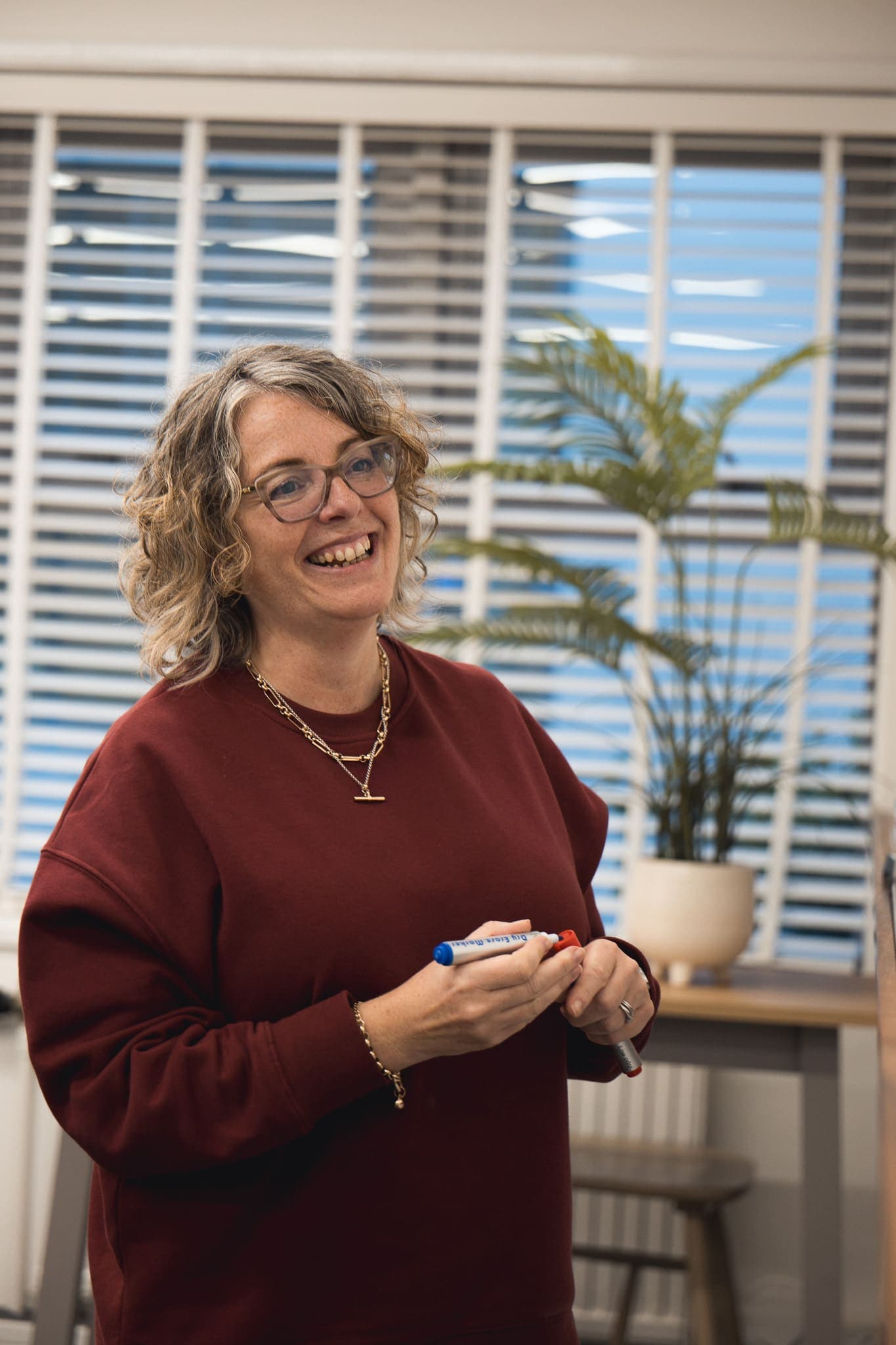 Smiling woman in glasses and a red sweatshirt holding whiteboard pens in a bright office with a potted plant.