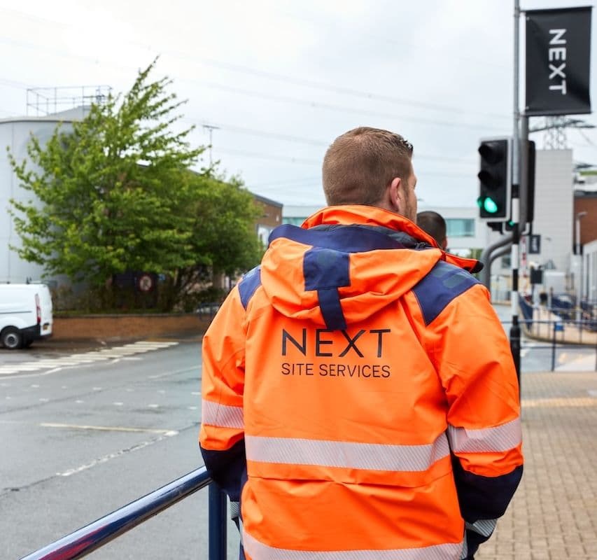Man in orange "NEXT Site Services" jacket standing near traffic lights outside an industrial building.