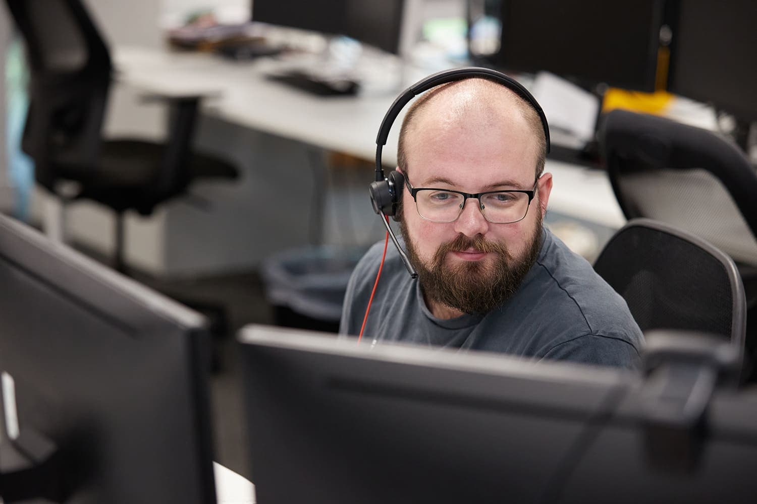 Bearded man wearing a headset looking at dual monitors in a modern office with empty chairs and desks.