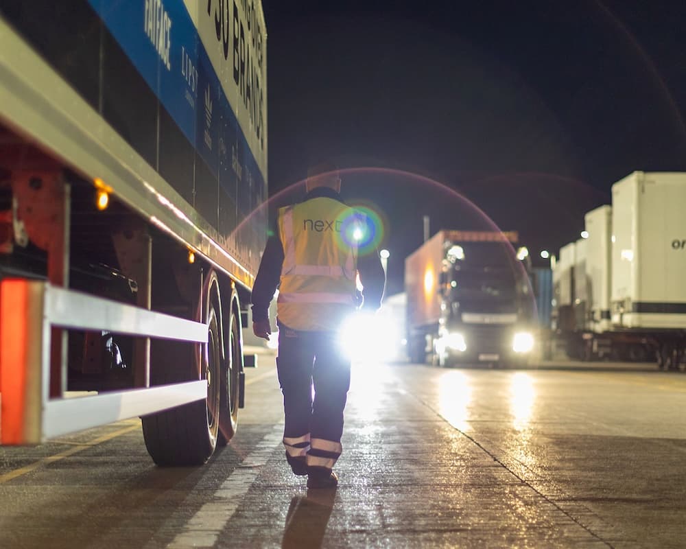 A logistics worker wearing a high-visibility "NEXT" vest walks beside a row of large trucks and trailers at night under bright lights, with a glow from headlights and lens flare in the background.