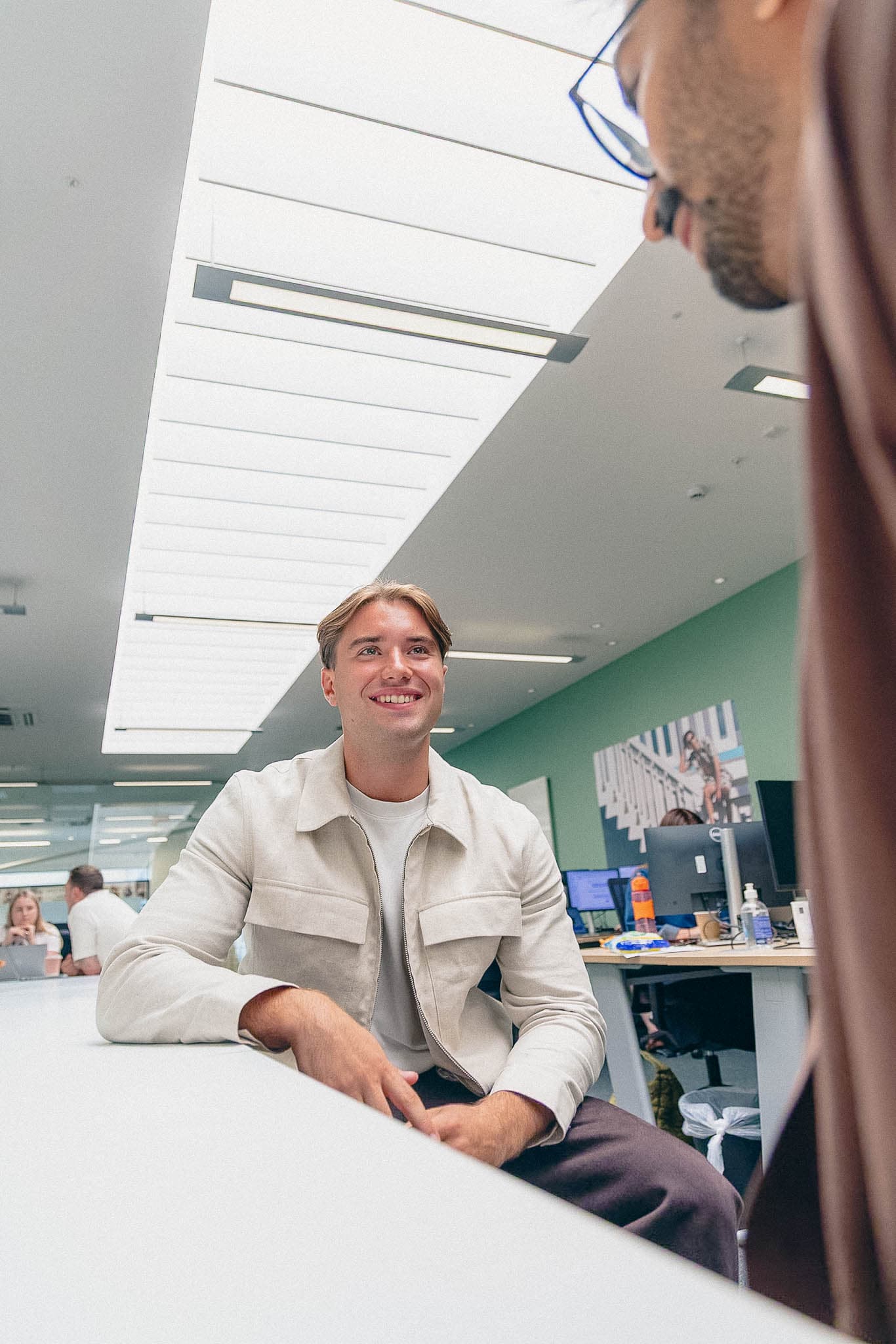 Man smiling while sitting at a desk in a bright, modern office space, speaking with a colleague out of focus in the foreground.