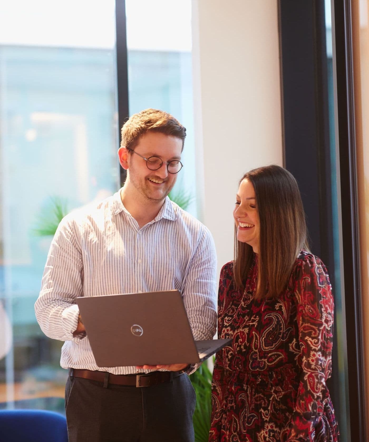 Two colleagues smiling while viewing a laptop screen together near large windows in a bright modern workspace.