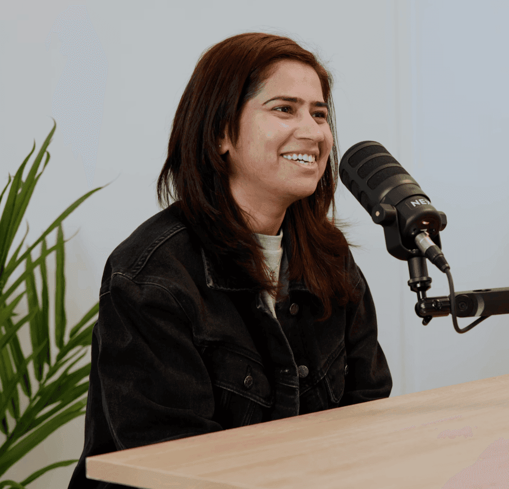 Smiling woman with dark hair wearing a black denim jacket, speaking into a black podcast microphone at a light wooden desk. A green plant is visible in the background.