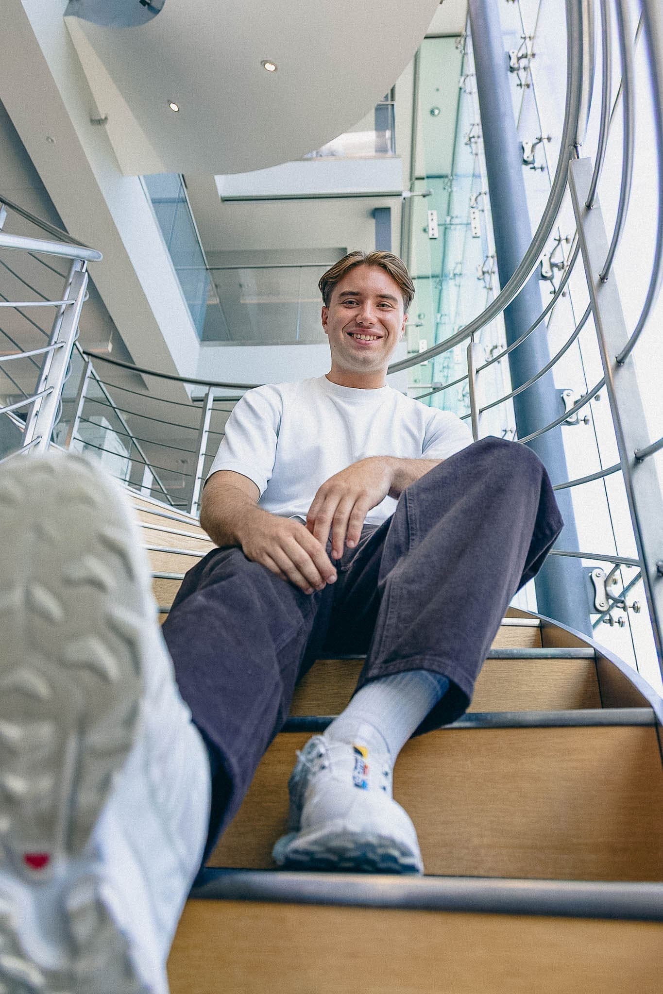Smiling male NEXT employee is sitting on indoor stairs in the office, wearing a casual white t-shirt and jeans.