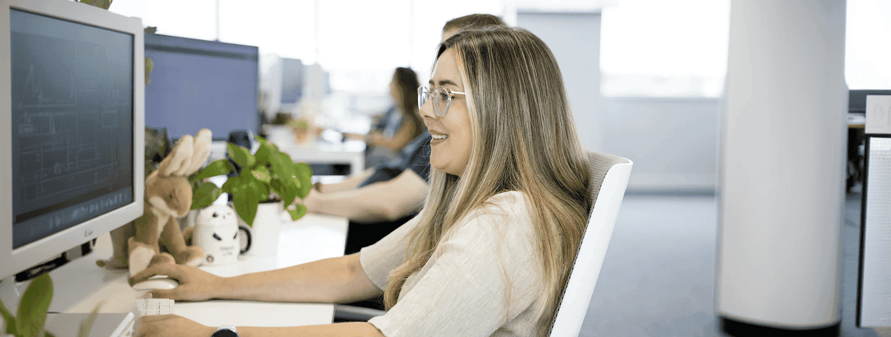 Woman sitting at a desk in an open-plan office, smiling while working at a computer, with plants and desk toys nearby.