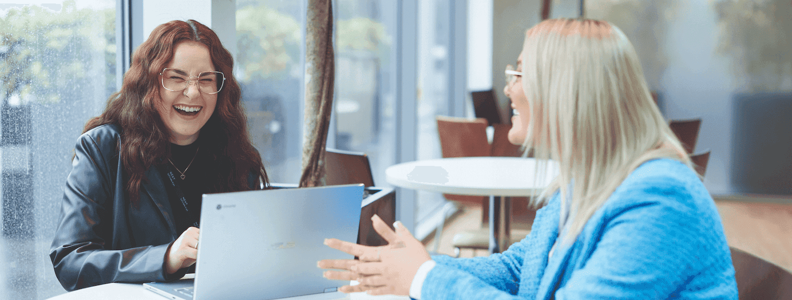 Two women sitting at a round table by large windows, laughing and talking while working on a laptop in a bright, modern office space.