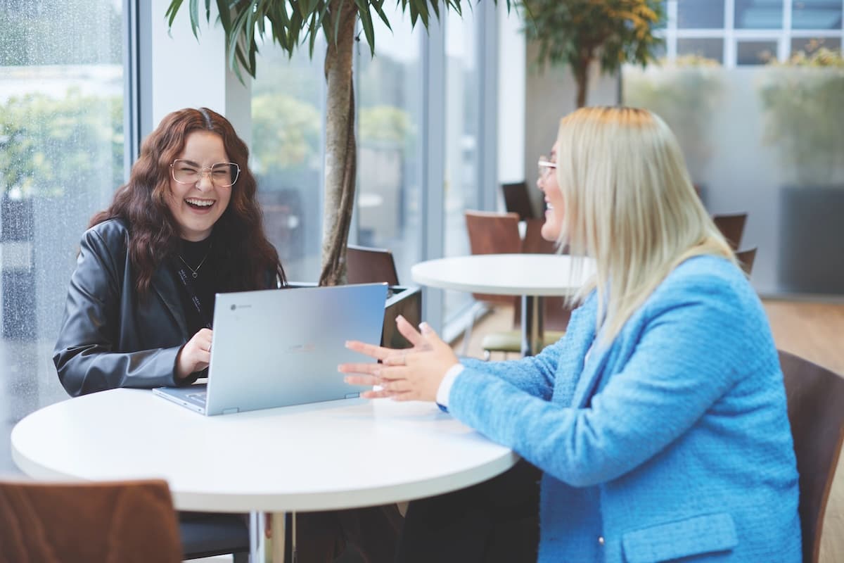 Two women sitting at a round table by large windows, laughing and talking while working on a laptop in a bright, modern office space.