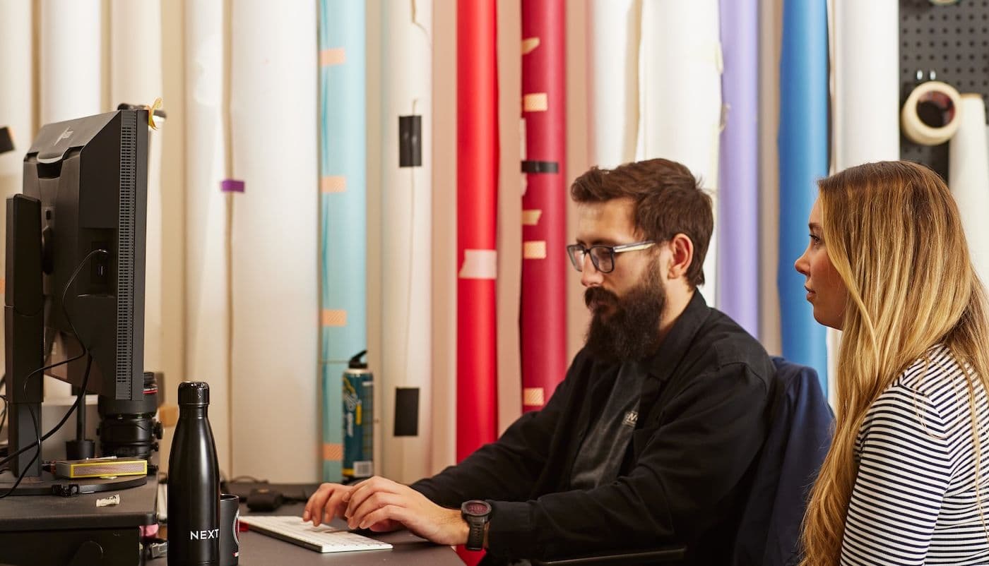 Man typing at a desk while a woman observes, with coloured backdrops and NEXT-branded items in the background.