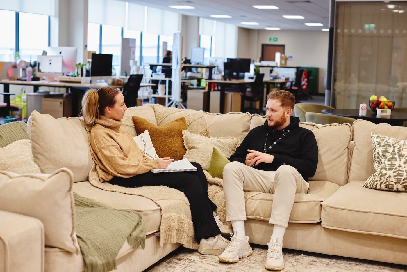 Two colleagues sitting on a sofa in a modern office, talking casually with a notepad open between them.