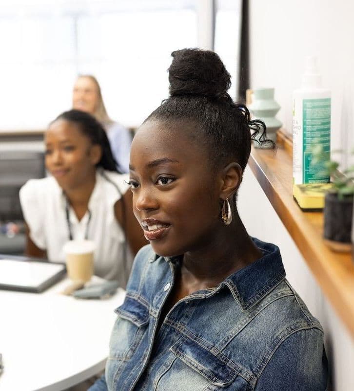 Group of women attending a meeting or workshop, seated around a table and listening attentively, with folders, drinks, and small plants on the table.