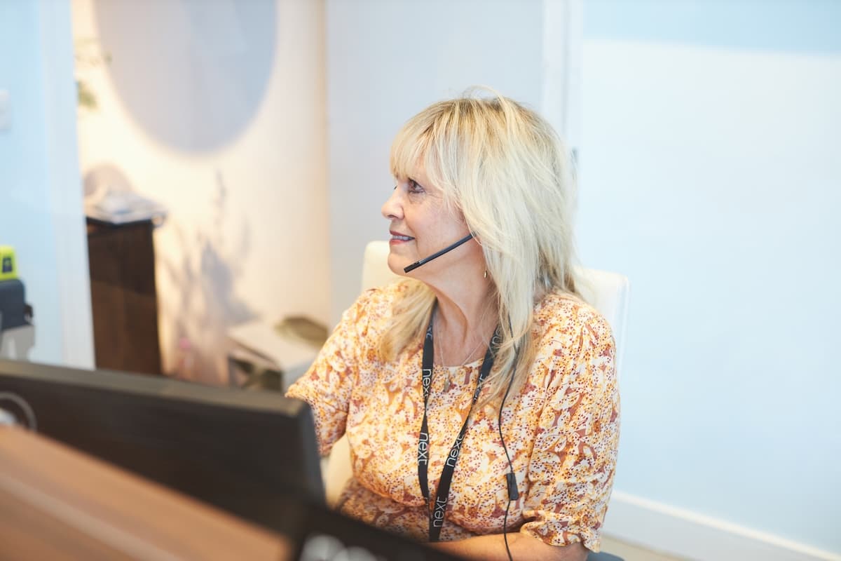 Smiling woman with a headset seated at a desk in a brightly lit reception area, wearing a floral top and a NEXT-branded lanyard.