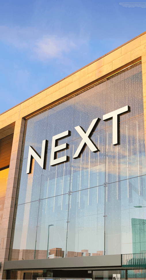 Exterior of a modern NEXT retail store with large glass windows and beige stone framing, reflecting the blue sky and clouds during sunset.