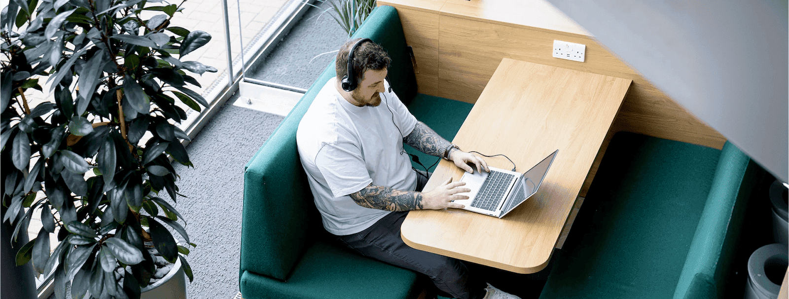 A man with tattoos, wearing a headset and white t-shirt, works on a laptop at a wooden booth in a modern, plant-filled office near a glass entrance.