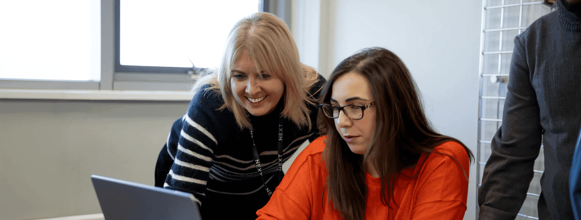 A woman in a striped jumper smiles as she leans over to assist a colleague in an orange jumper, who is working on a laptop at a desk in an office setting.