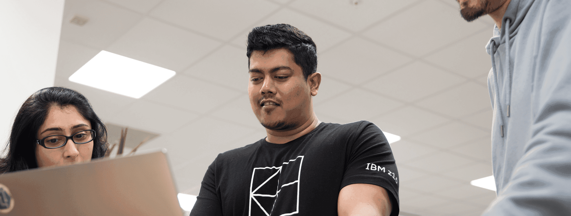 Man in a black top works at a laptop in the office, looking down and smiling slightly. 