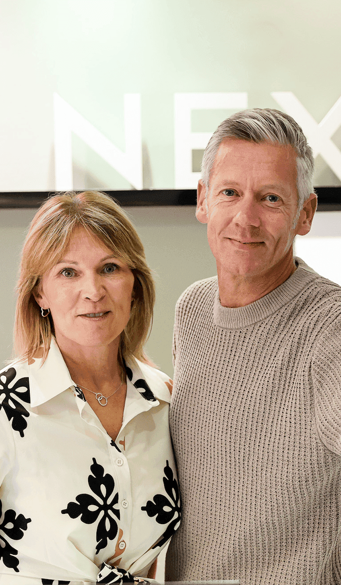 Two smiling colleagues standing in front of a reception desk with the NEXT logo displayed behind them.