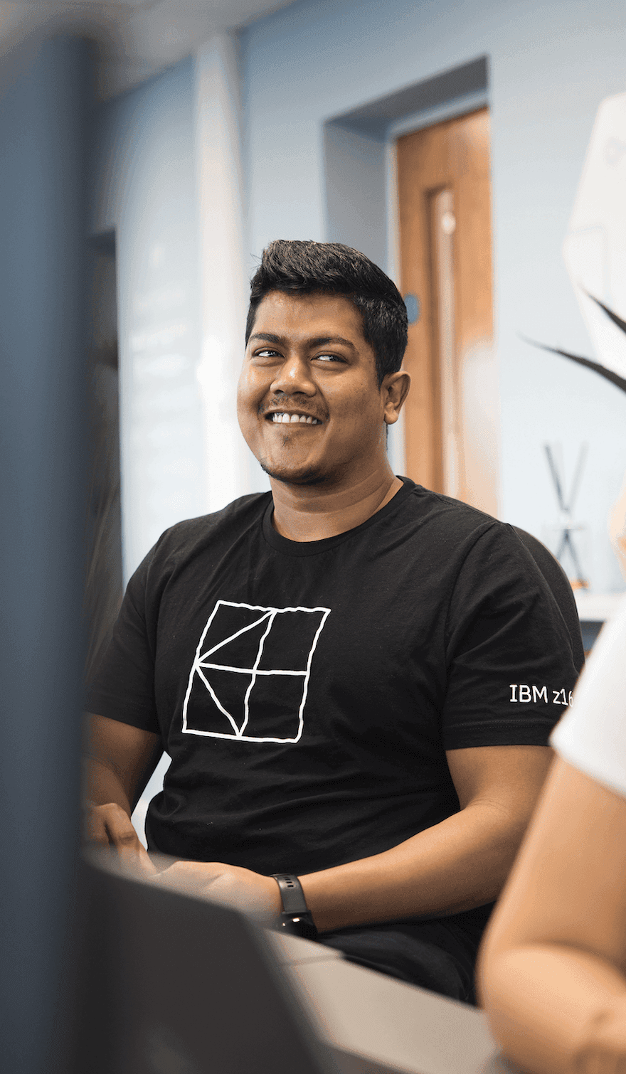 Smiling man wearing a black IBM t-shirt, seated in an office with a blue wall and a wooden door in the background.