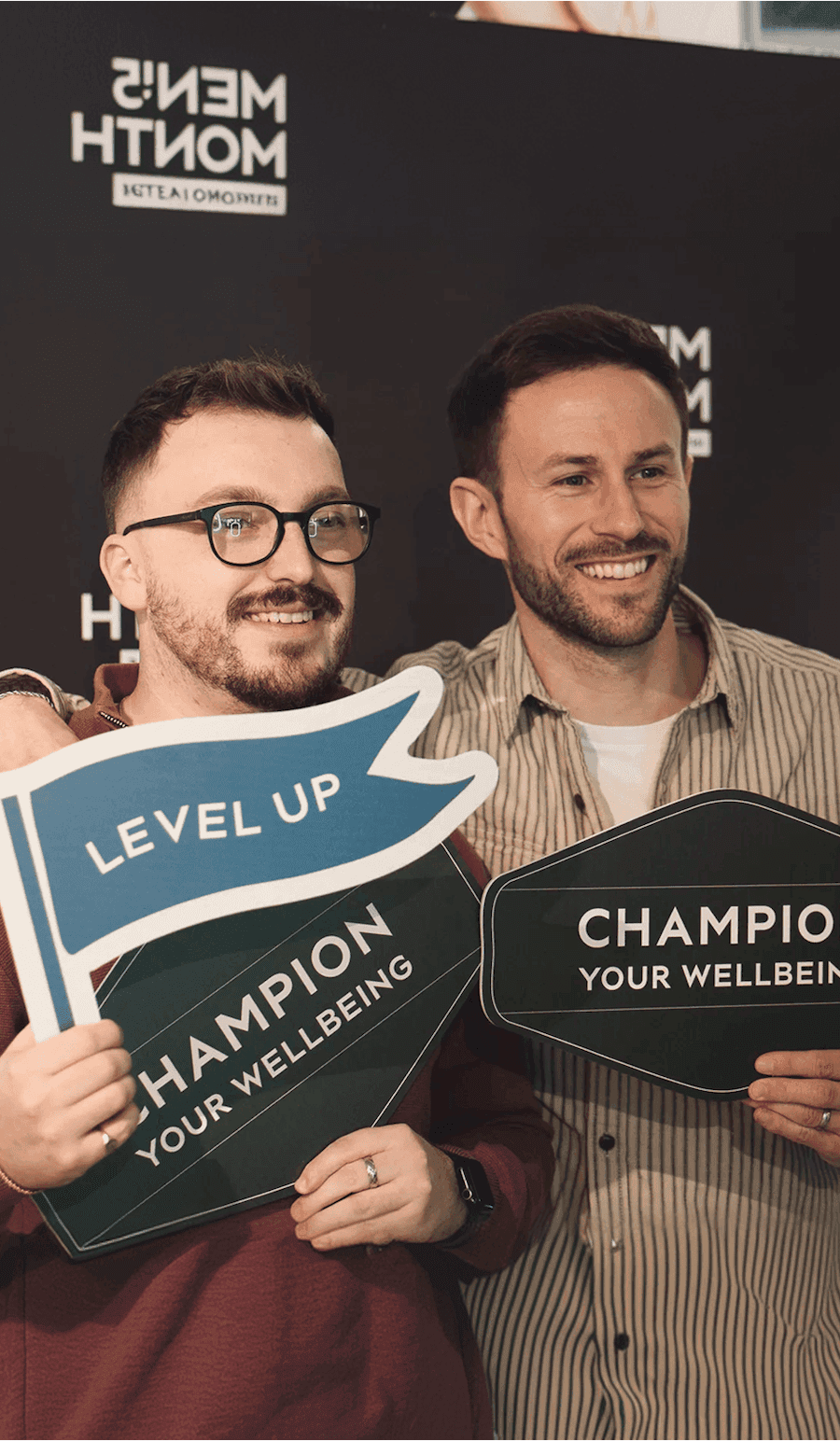 Two men standing next to one another at a Men’s Month event, holding up signs that say “LEVEL UP” and “CHAMPION YOUR WELLBEING.”