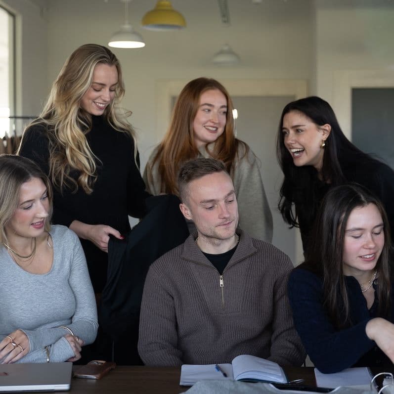 A group of young professionals collaborate around a table, smiling and discussing fabric samples and notebooks in a modern fashion studio setting.