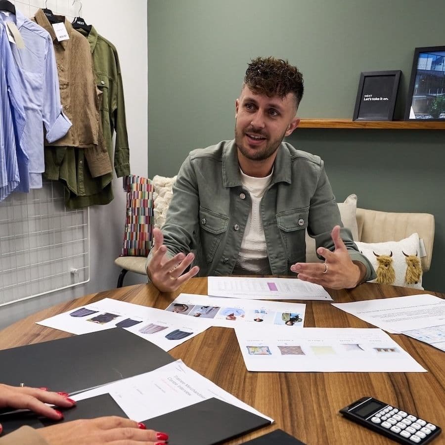 A man sits at a table with fashion documents, fabric samples, and a notebook labelled "EARLY CAREERS NEXT," engaged in discussion with colleagues in a workspace decorated with hanging clothes.