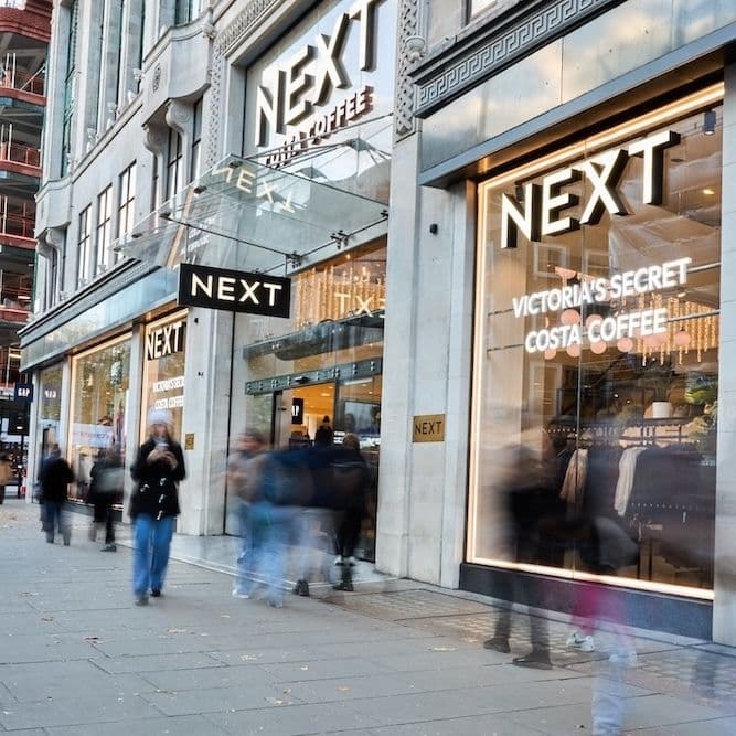 Street view of a NEXT flagship store with illuminated signage and window displays featuring Victoria’s Secret and Costa Coffee. Pedestrians blur past, suggesting a busy shopping area.