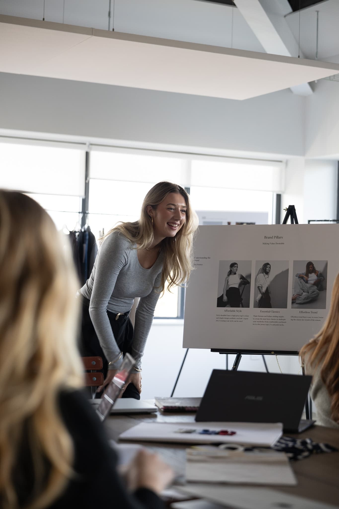 A smiling woman in a grey top presents to a group, standing beside a display board showing brand pillars and fashion photography in a bright, modern office.