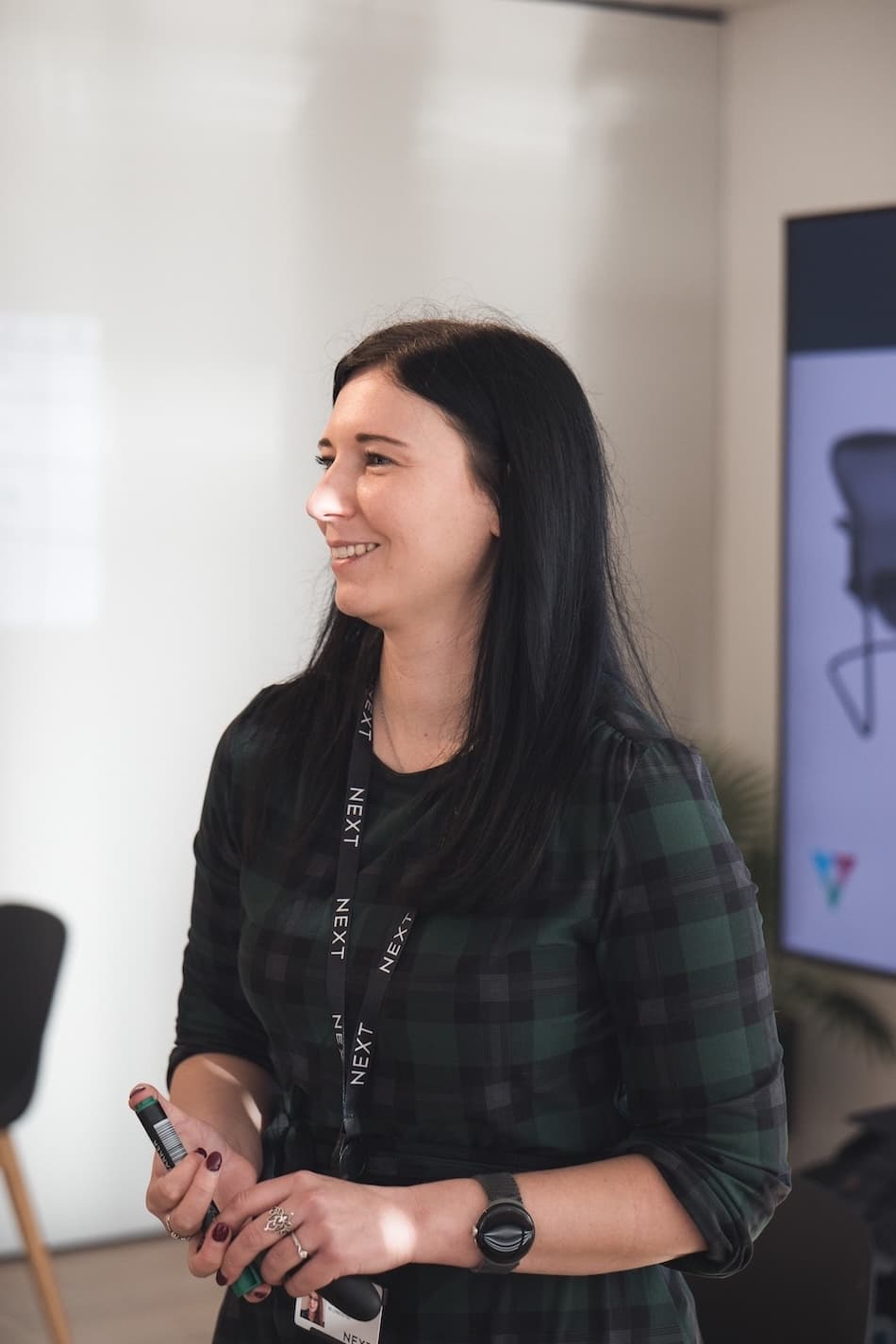 A woman wearing a NEXT-branded lanyard and a green plaid dress smiles while speaking in a well-lit meeting space, holding a marker in her hand.