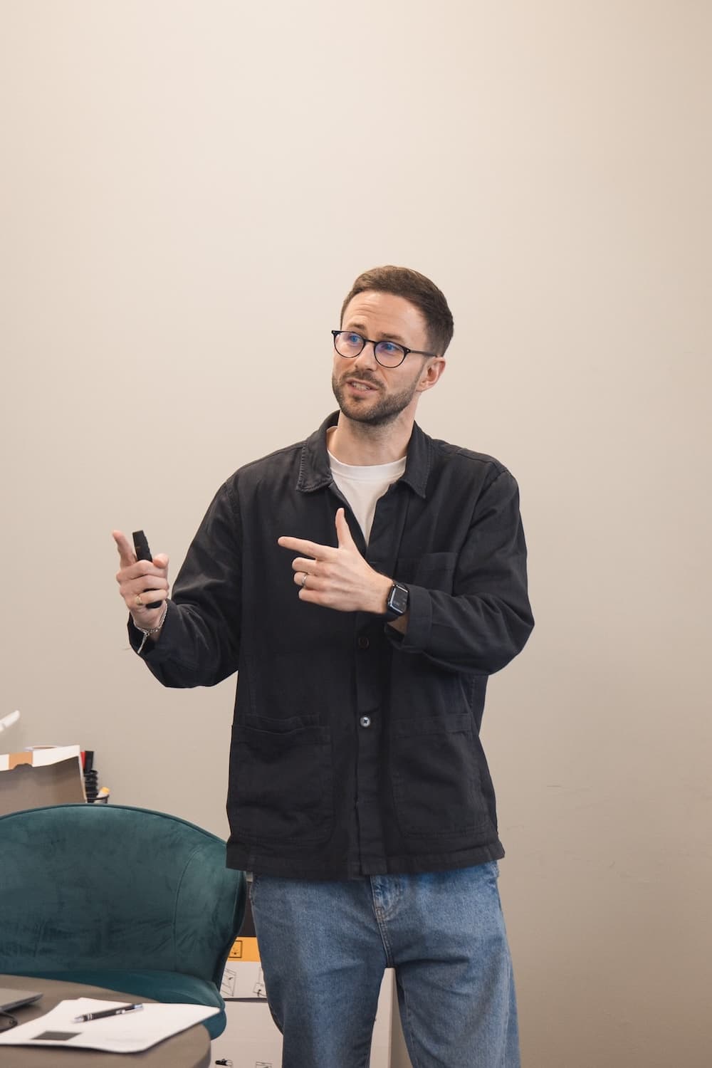 A man in glasses and a dark jacket gestures while presenting in front of a light wall. He holds a remote and wears a smartwatch, standing near a table and a green chair in a workspace.