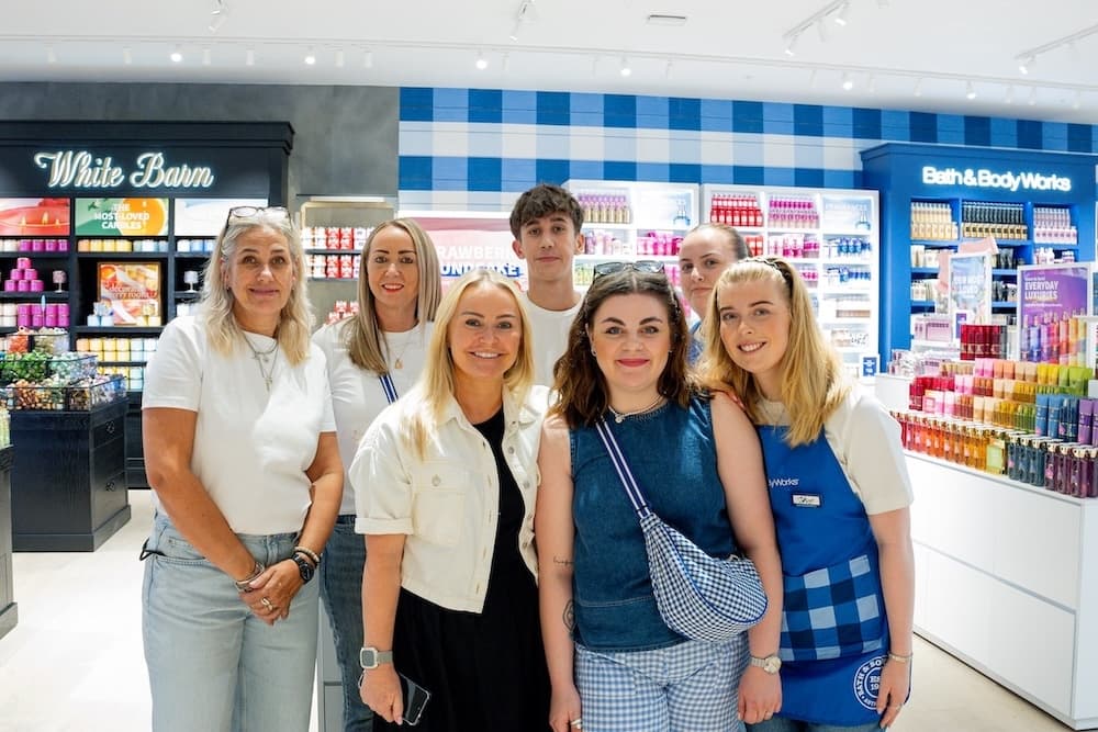 A group of smiling employees and team members stands together inside a brightly lit Bath & Body Works store, surrounded by colourful displays of candles, lotions, and fragrances.