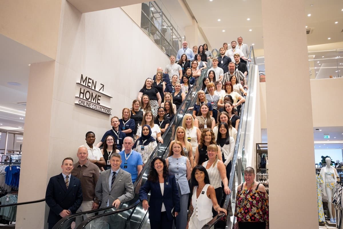 A large group of employees poses on both sides of an escalator inside a department store. A sign reading "MEN ↑ HOME ONLINE COLLECTIONS" is visible on the wall, and the atmosphere is bright and professional.