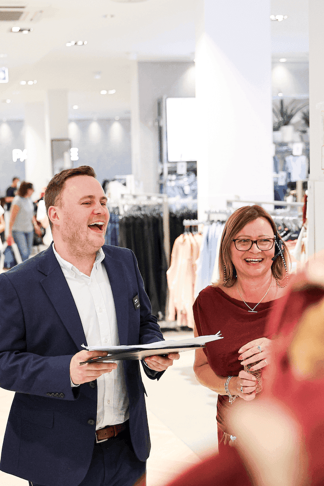 Man in a navy suit and woman in a headset laughing together while holding papers in a busy clothing store.