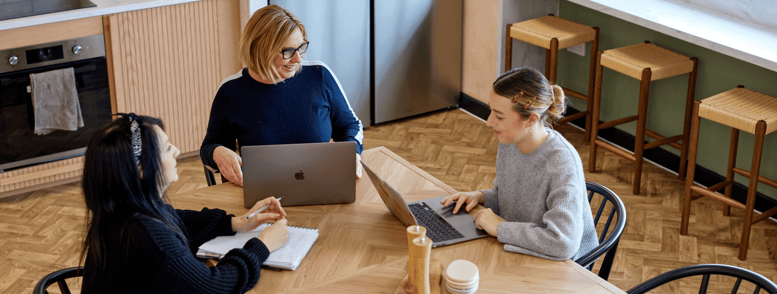 Three women seated around a wooden kitchen-style table, engaged in discussion while using laptops and taking notes in a cosy office environment.