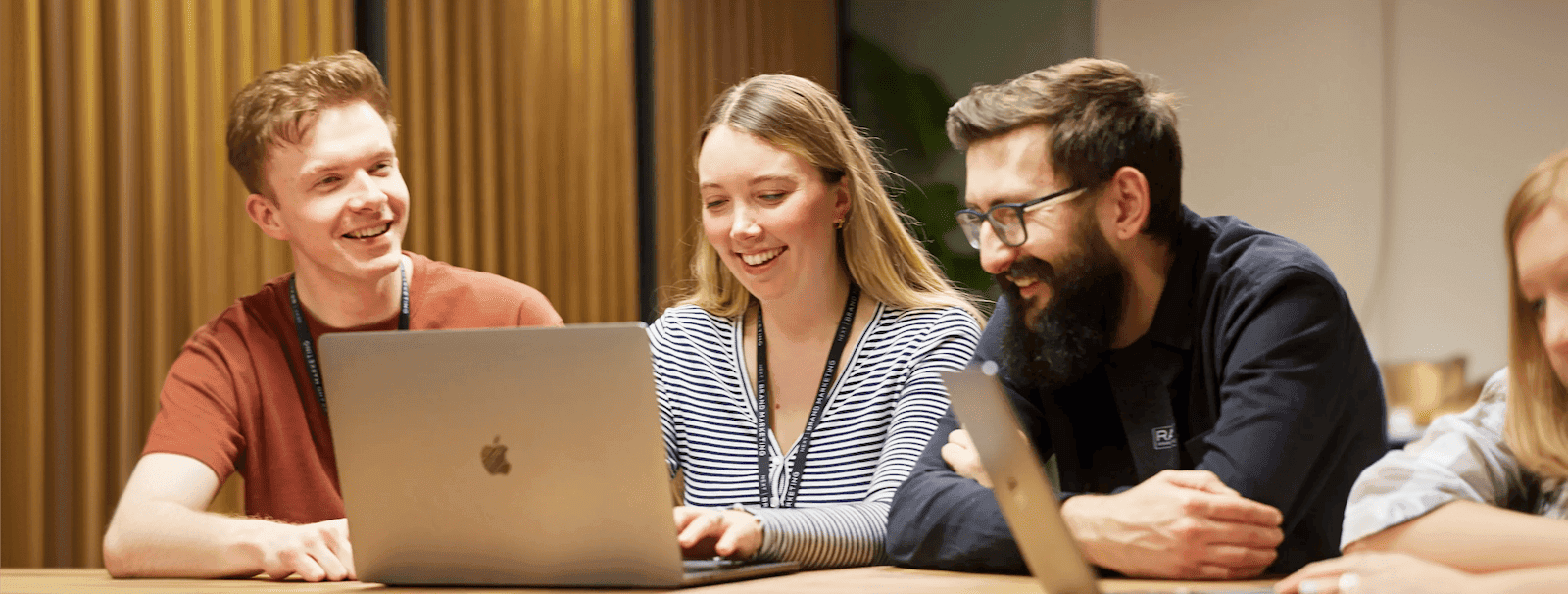 Three colleagues smiling and working together at a table, looking at laptops, with visible NEXT branded lanyards and a modern office setting.