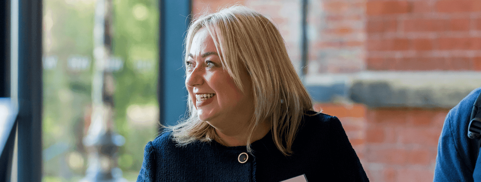 Smiling woman with blonde hair looking to the side, indoors near large windows with natural light and a brick wall background.