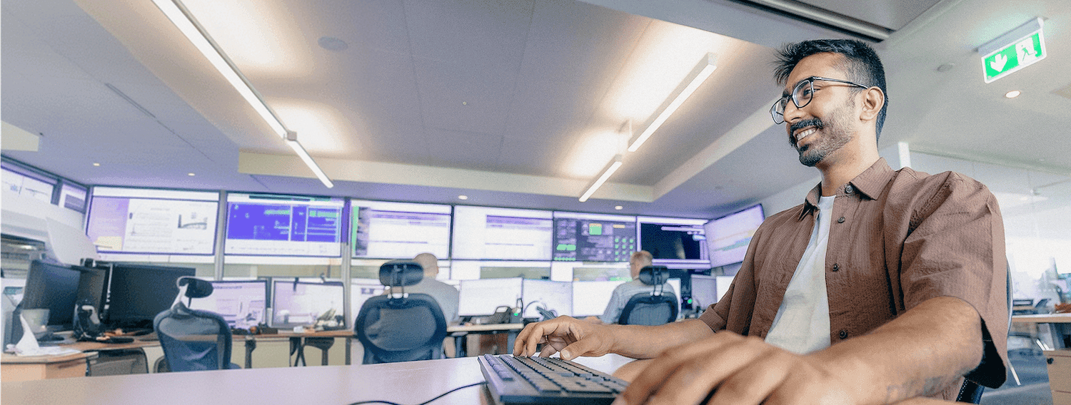 Smiling man in glasses using a computer in a modern operations or control room, with multiple screens displaying data in the background.