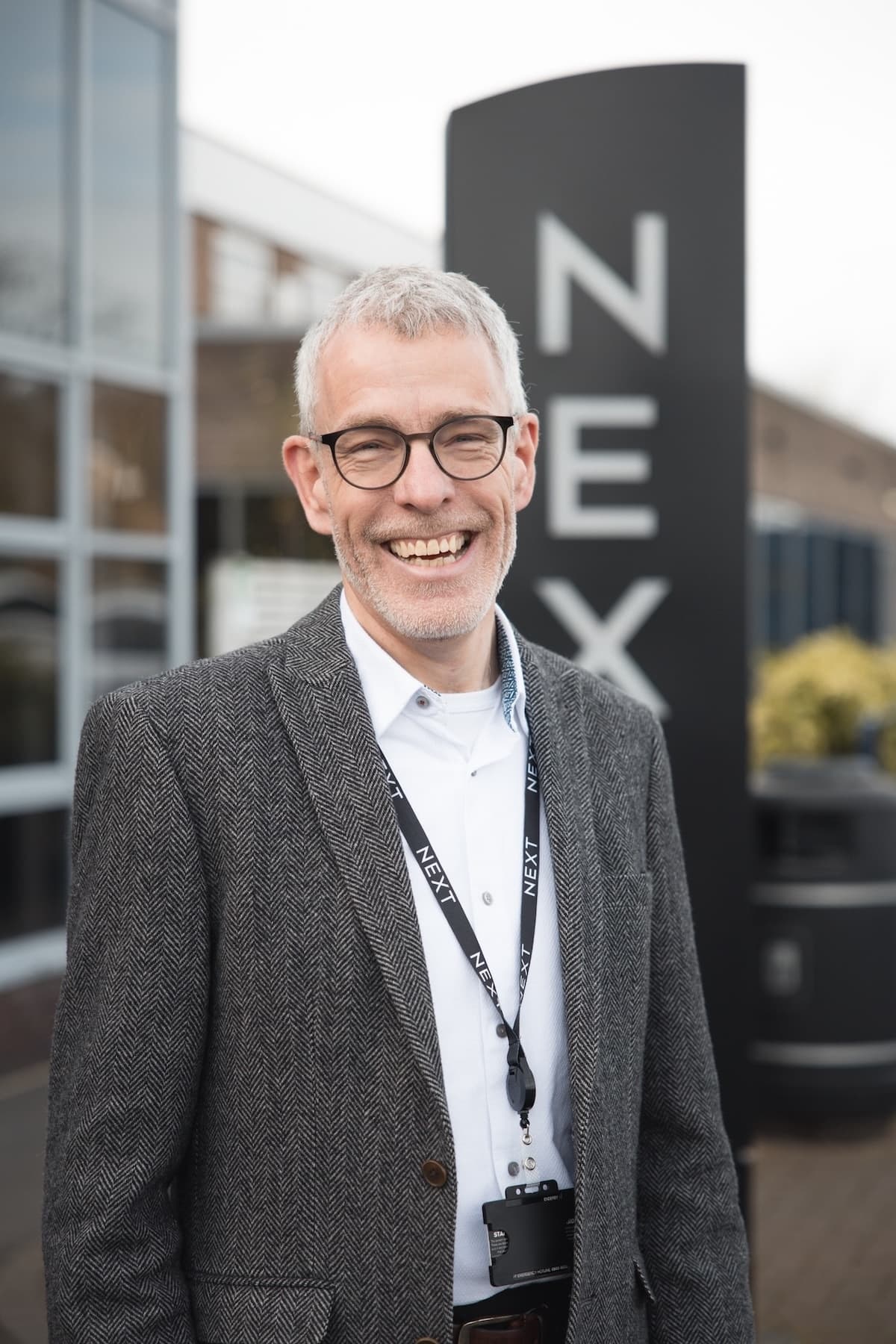Smiling man wearing glasses, a grey blazer, and a lanyard with the NEXT logo, standing outside a NEXT office building with a tall black NEXT sign in the background.