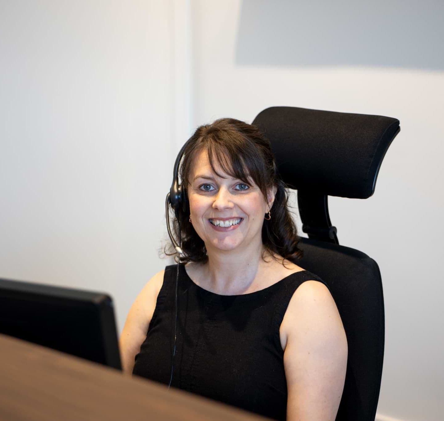 Smiling female NEXT employee with short brown hair, sitting at desk wearing a headset with a microphone. 