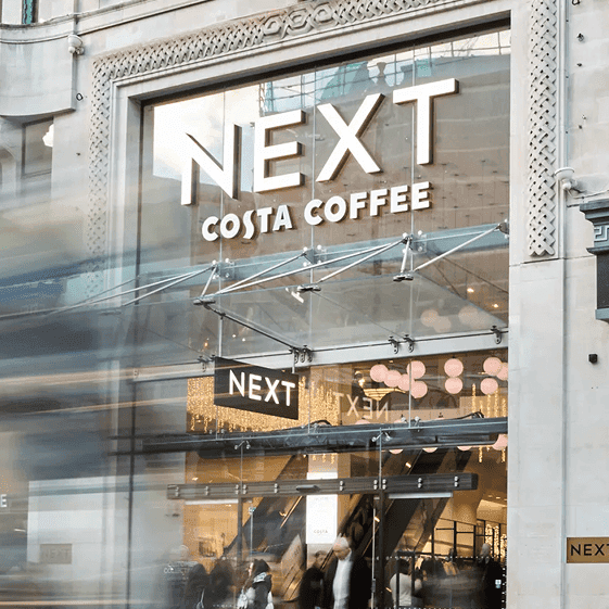 Street-level view of a NEXT retail store featuring signage for Costa Coffee and Victoria’s Secret, with a blurred double-decker bus passing in front.
