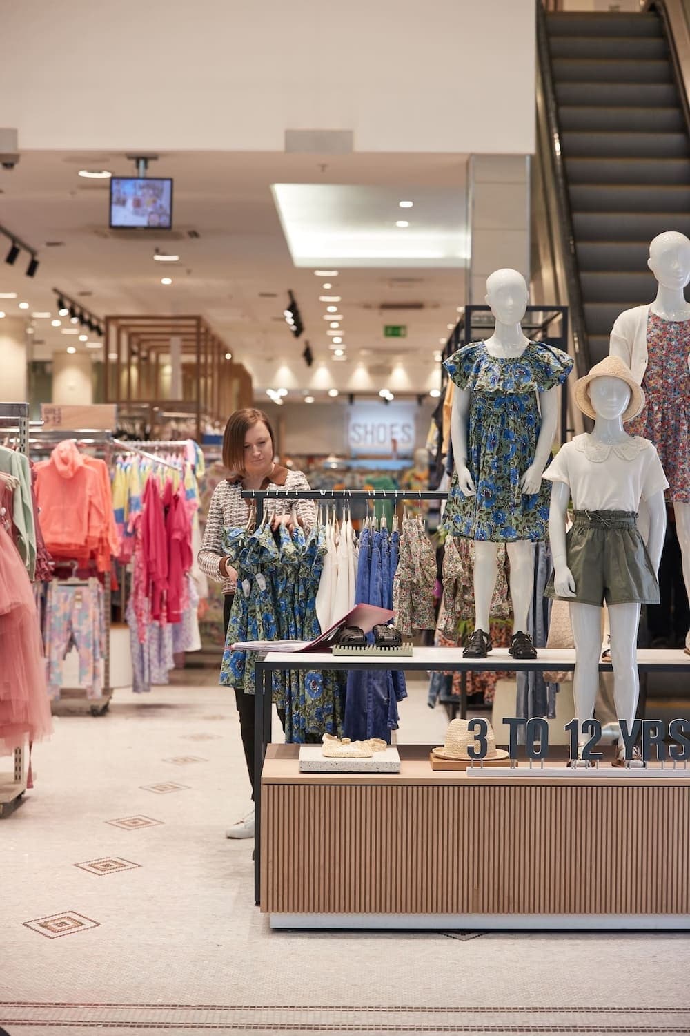 A retail employee arranges children’s clothing on a rack in a brightly lit NEXT store, surrounded by colourful displays and mannequins.