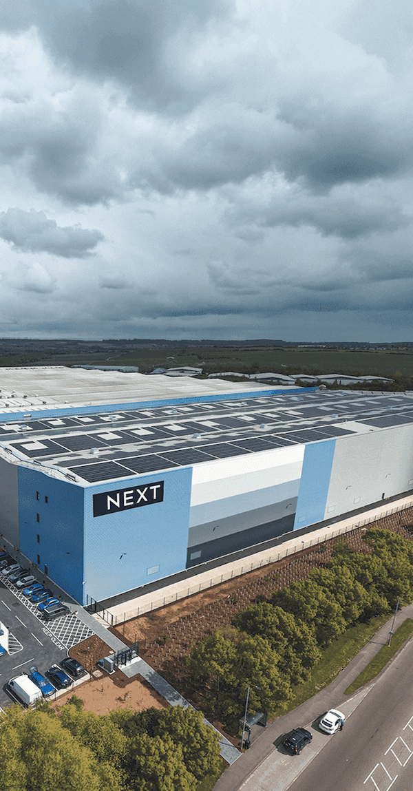 Aerial view of a large NEXT distribution centre with solar panels on the roof, extensive parking, and surrounding roads under a partly cloudy sky.