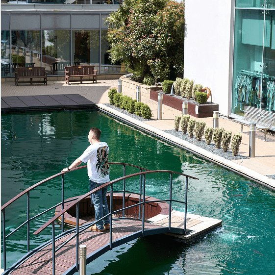 A man stands on a curved footbridge over a green reflecting pool outside a modern office building, surrounded by neatly trimmed bushes and wooden benches.