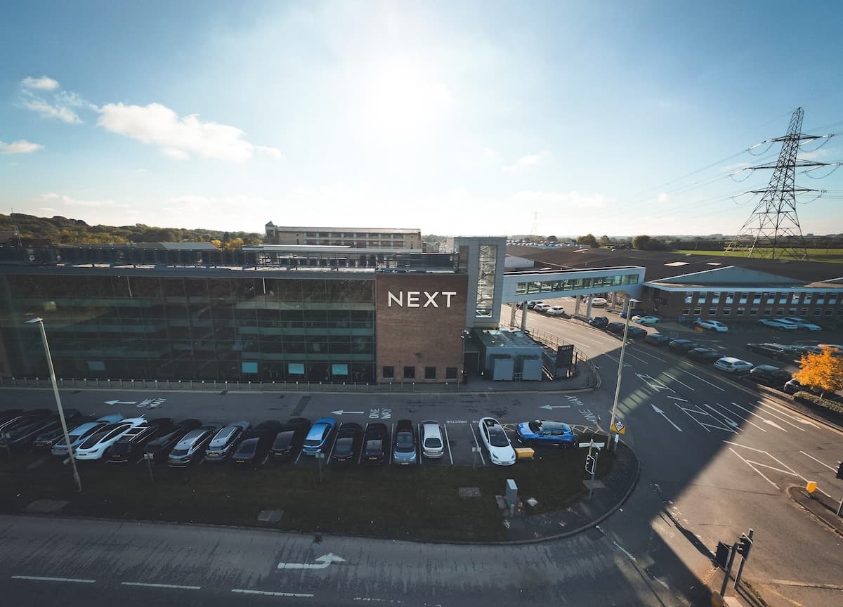 Exterior view of a NEXT office building with modern glass and brick architecture, adjacent to a road and connected by a glass walkway.