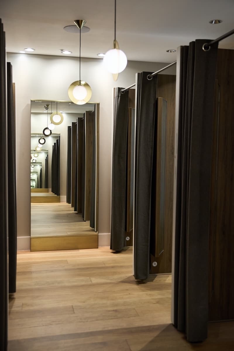 Modern retail fitting room corridor with wooden flooring, dark curtains, and stylish pendant lighting, reflected infinitely in a large mirror at the end.