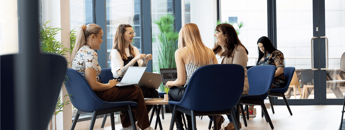 Four women sitting in a modern, brightly lit lounge area with laptops open, engaged in conversation, while another woman works in the background.