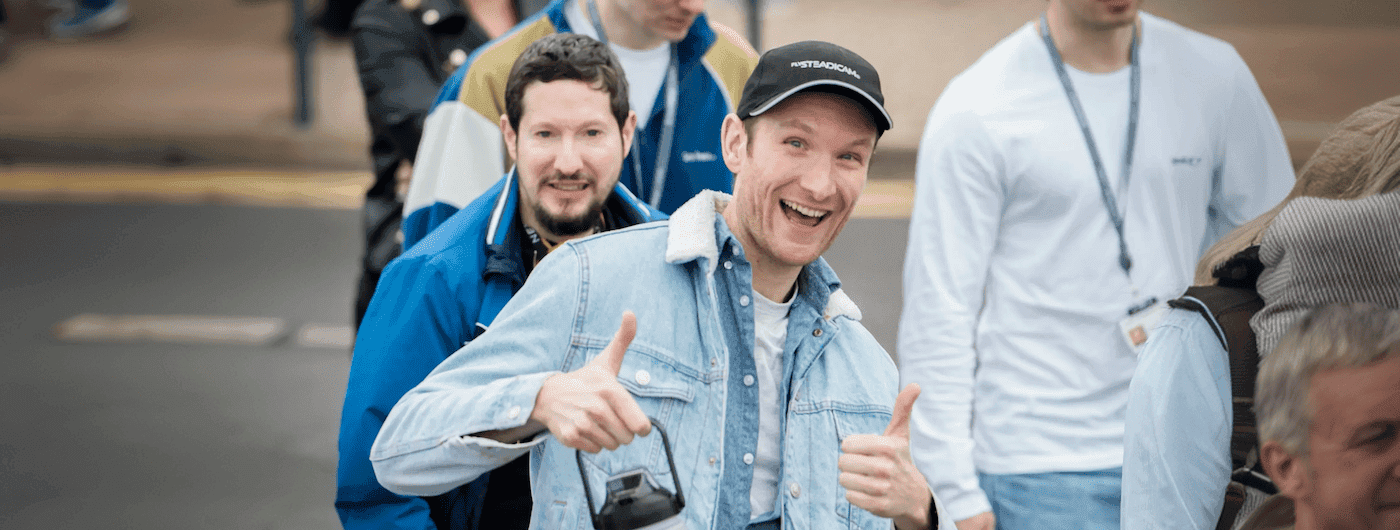 Man in a denim jacket smiling and giving two thumbs up while walking outdoors with a group of people, some wearing lanyards.