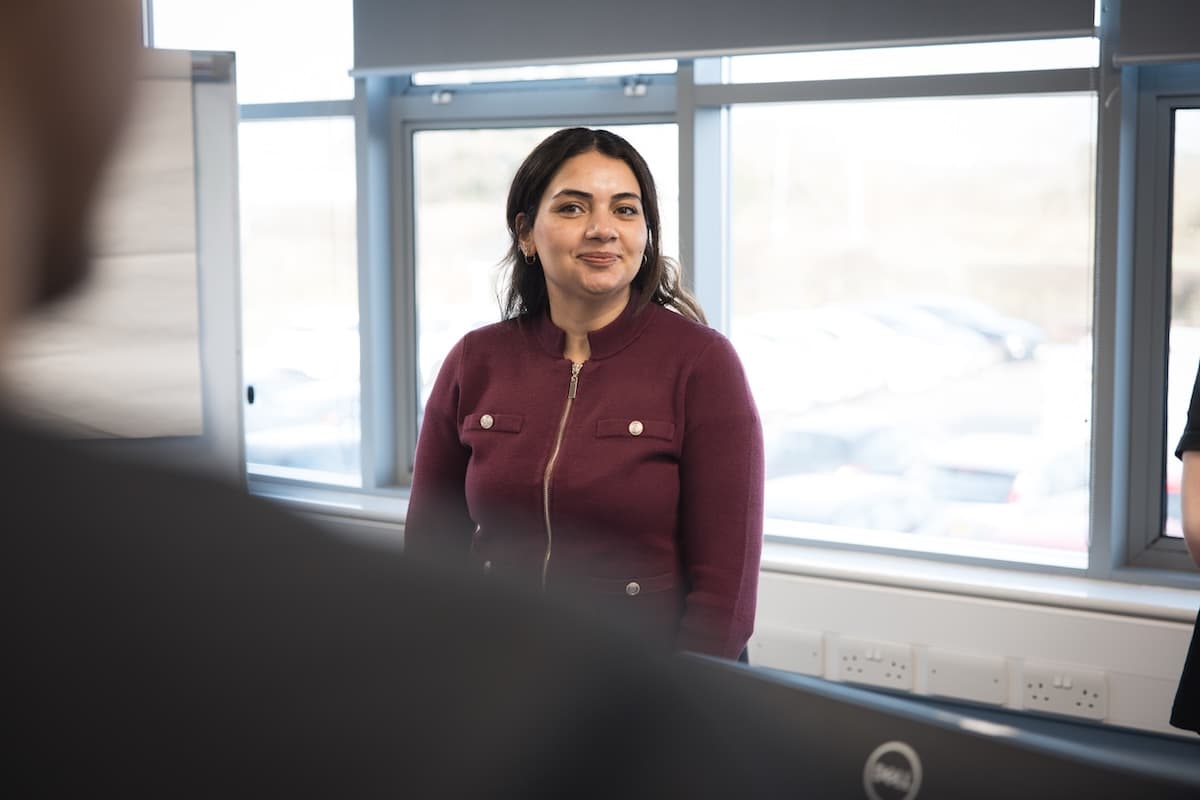 A woman in a burgundy jacket stands in front of row of windows, smiling, during a presentation or meeting in a bright office space with large windows.