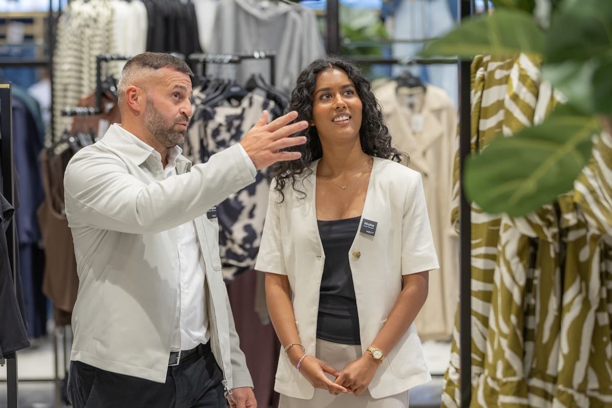 Two retail employees stand among clothing racks having a discussion. The man gestures while the woman, wearing a NEXT name badge, listens attentively and smiles.