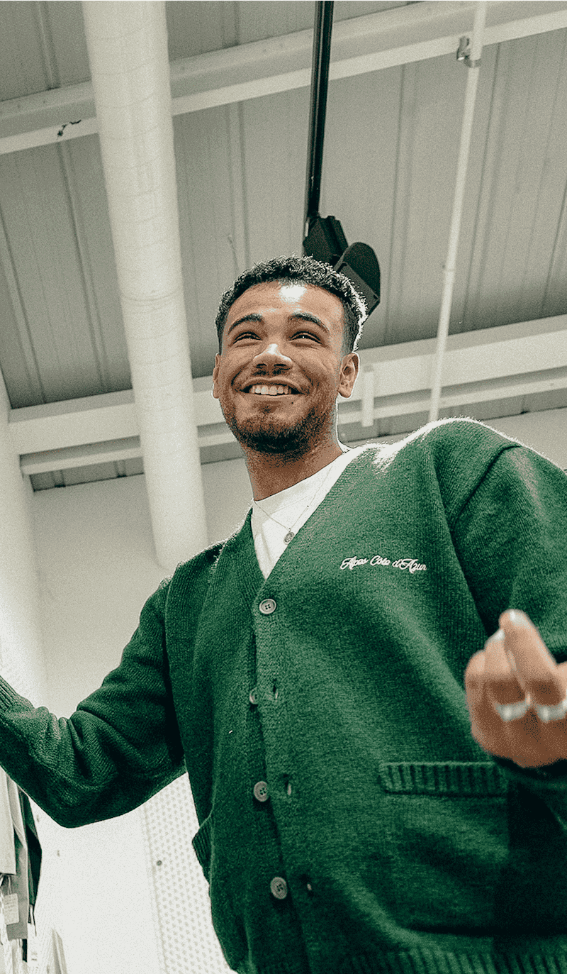 Smiling person in a green cardigan standing next to a wall display of hanging clothing items in a modern showroom.