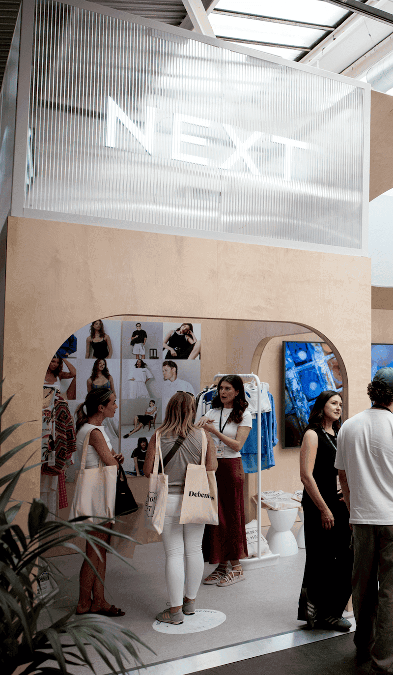 Group of people interacting at a NEXT exhibition booth with mannequins, screens, and fashion displays in a bright indoor space.